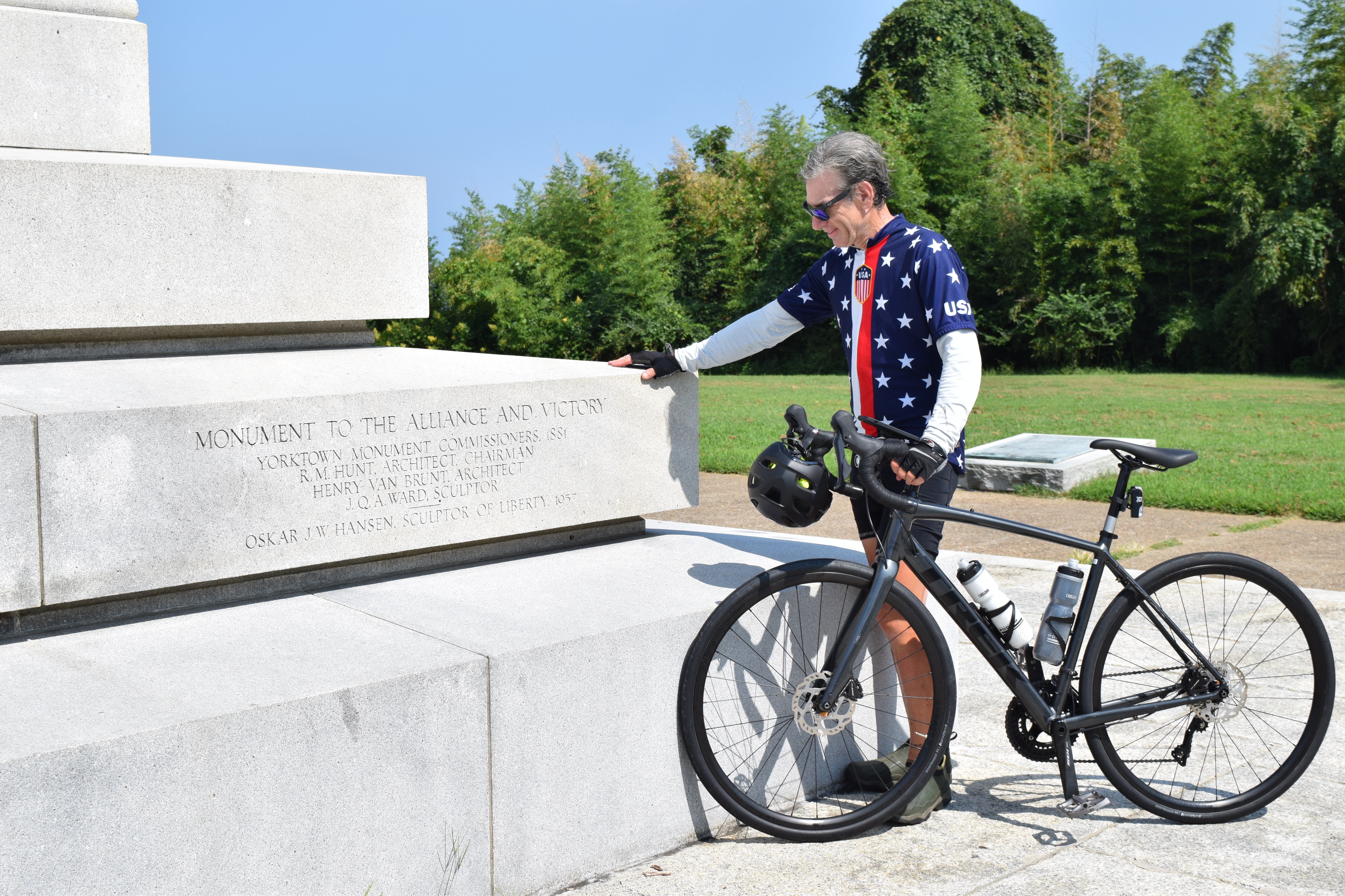 Sal Lilienthal stands next to the white stone Yorktown Monument with his bike wearing an American flag windbreaker
