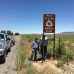 Two men pose under a sign reading "Pony Epress Historic Rout" next to a highway
