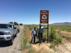 Two men pose under a sign reading "Pony Epress Historic Rout" next to a highway